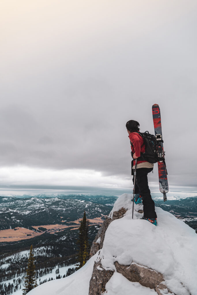 A snowboarder stands at the top of a narrow peak and surveys the landscape below.