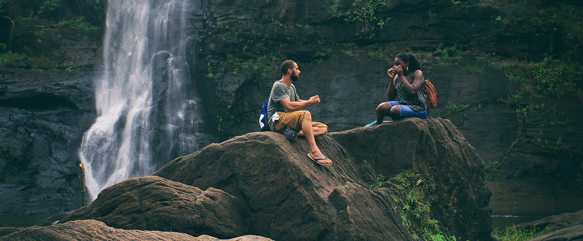 Two hikers chat on tall rocks in front of a waterfall.