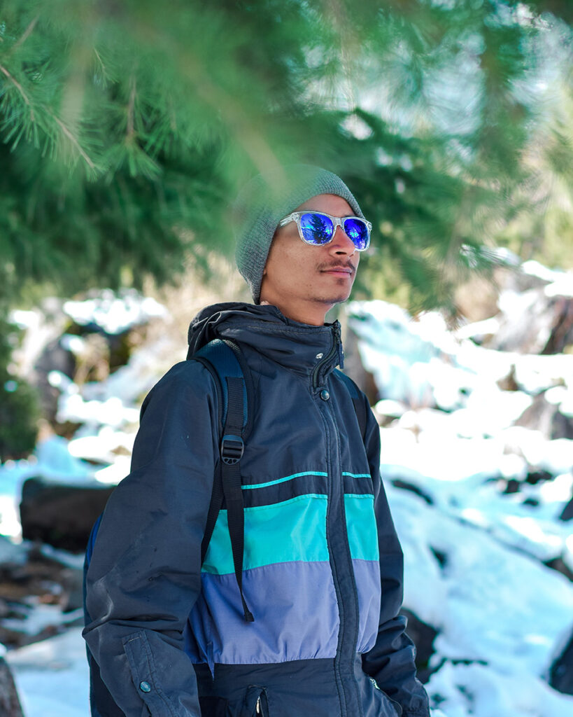 A hiker in sunglasses gazes out from under a pine tree.