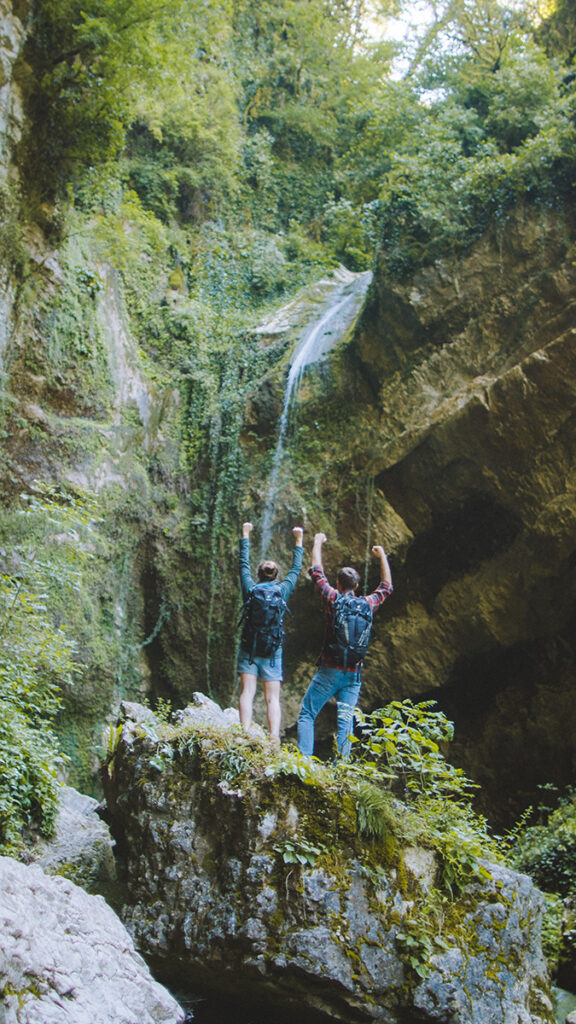 Two hikers raise their arms in celebration in front of a waterfall.