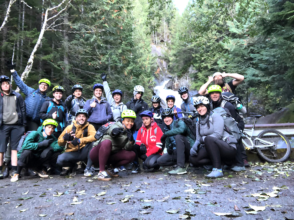 About 18 students on a bike ride, posing on a bridge over a river in the forest.