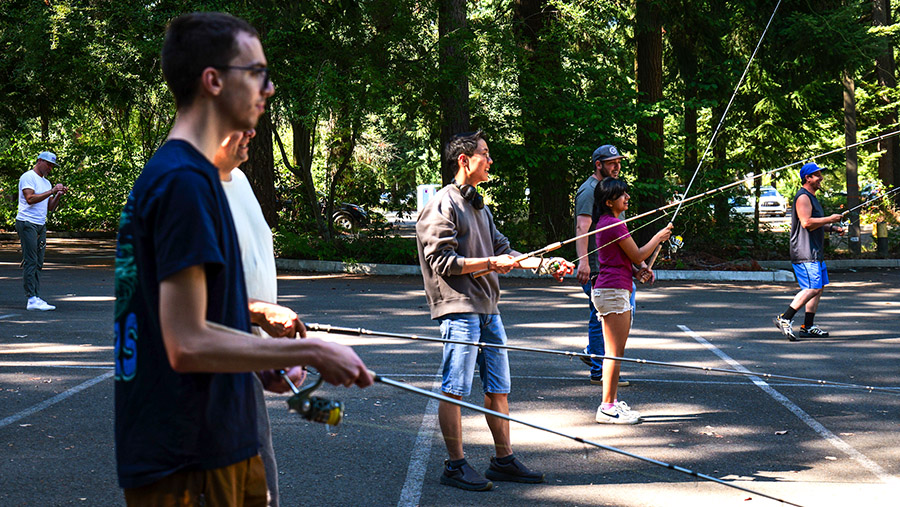 Students in summer attire practicing their flycasting techniques in the parking lot with the trees casting bright shadows.