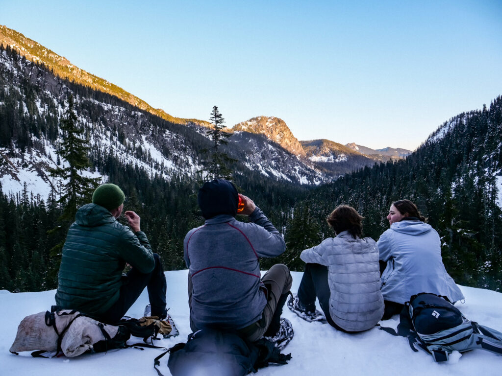 Tucked above Source Lake, snowshoers on a wintry day sit on the snow and watch the fading sun peeking through the mountains.