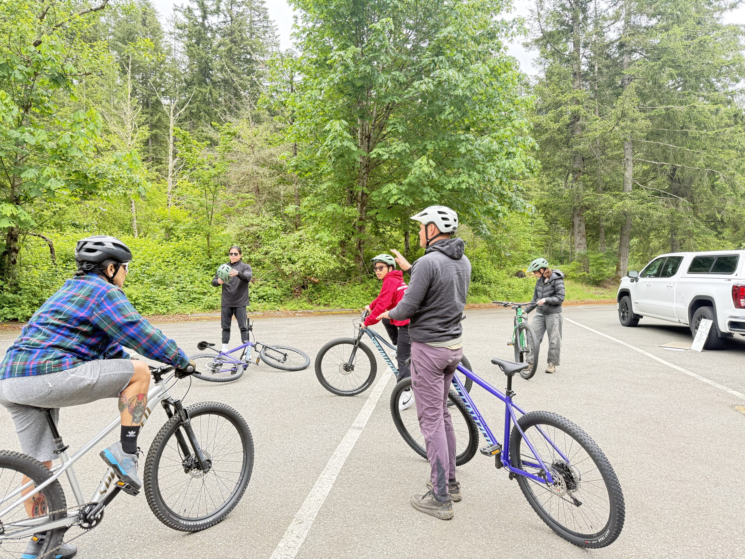Students and their instructor in helmets, mounting their bikes at the trailhead to Olallie State Park, surrounded by huge hardwood and conifer trees.