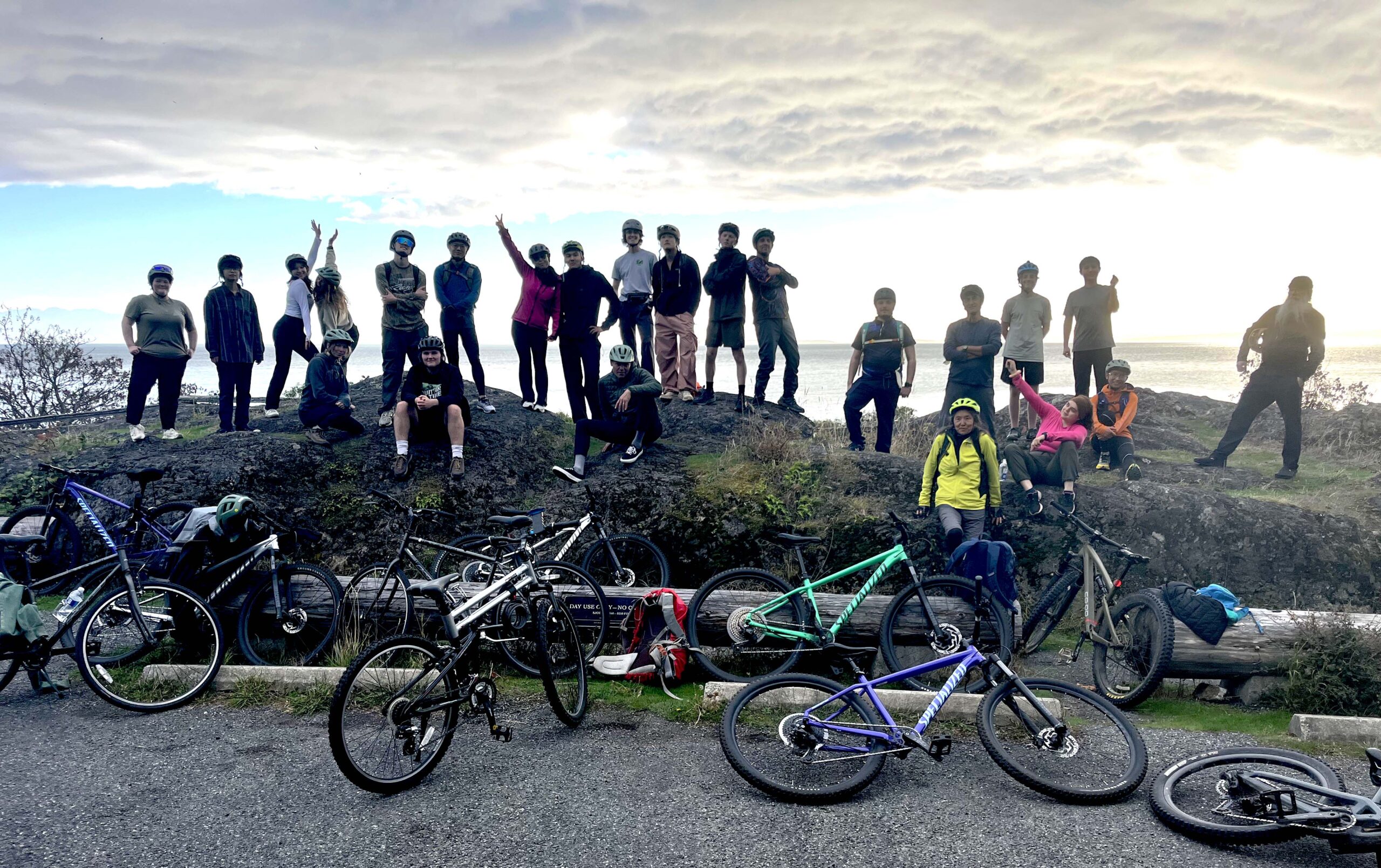 A large cadre of students standing on a berm silhouetted in the setting sun with Haro Strait in the background and bikes leaning in front.
