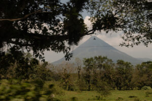A hint of Arenal volcano in the distance under a hazy sky from behind the dark tree leaves.