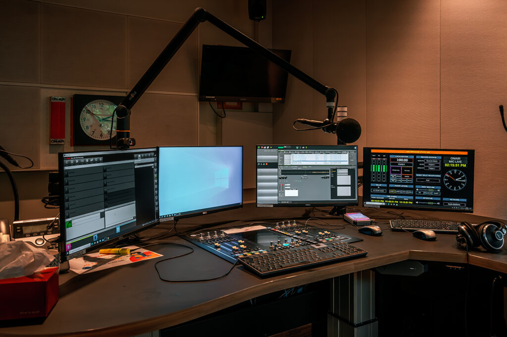 The inside of a radio broadcast booth shows various computers and sound engineering technology.