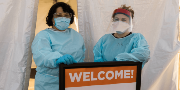 Nurses in scrubs and masks stand by a welcome sign