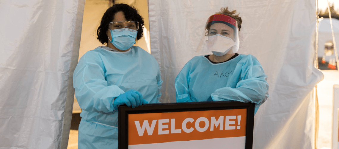 Nurses in scrubs and masks stand by a welcome sign