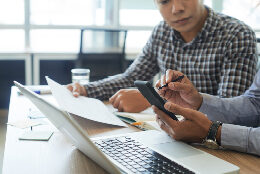 Two people work on a phone, laptop, and with documents while they discuss.