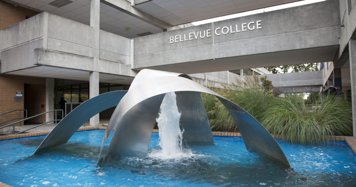 A large outdoor fountain with multiple silver arches and a single water spout in the center. The second level walkway is in the background with the Bellevue College sign in the center.