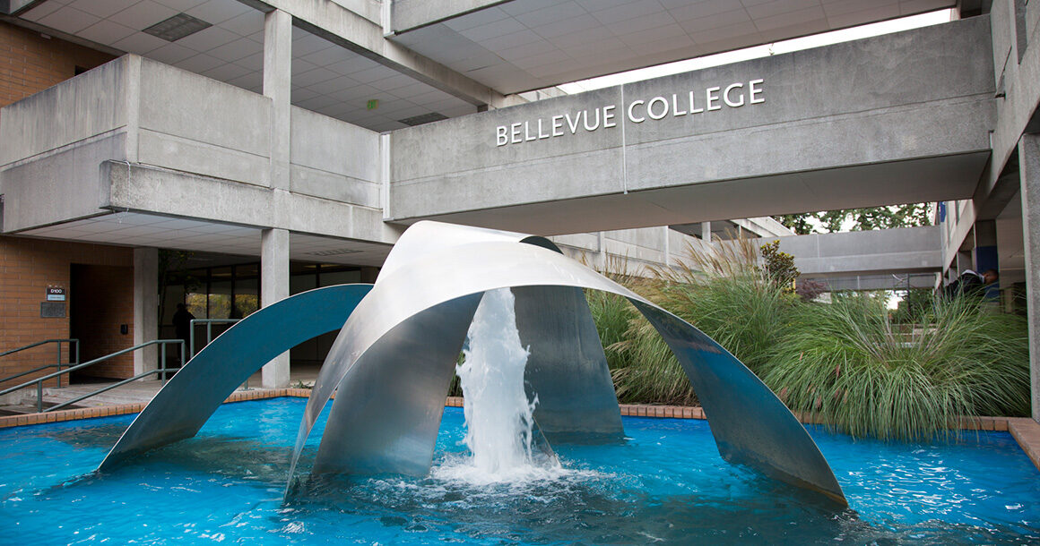 A large outdoor fountain with multiple silver arches and a single water spout in the center. The second level walkway is in the background with the Bellevue College sign in the center.