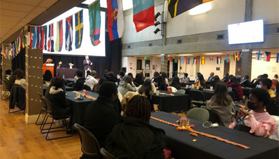 Students and staff gather in the cafeteria, where flags from many countries hang above the entry ways. Tables with black tablecloths and autumnal decorations are spaced with four seats each: two on either side. Seated individuals look toward a stage where a person in a suit jacket addresses the crowd.