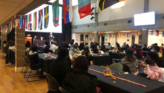 Students and staff gather in the cafeteria, where flags from many countries hang above the entry ways. Tables with black tablecloths and autumnal decorations are spaced with four seats each: two on either side. Seated individuals look toward a stage where a person in a suit jacket addresses the crowd.