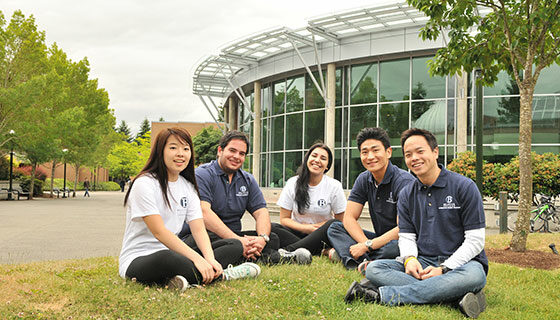 Students sitting on the grass in a circle smiling in front of the C building.