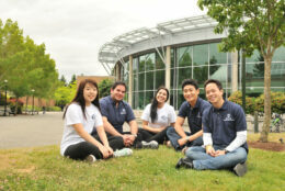 Students sitting on the grass in a circle smiling in front of the C building.