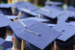 Students wearing graduation caps sit attentively at commencement, as seen from behind.