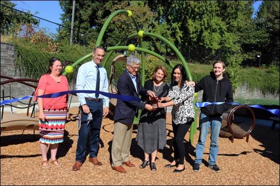 A group gathers beside play equipment to cut a ribbon stretched across the sunny ELC playground.