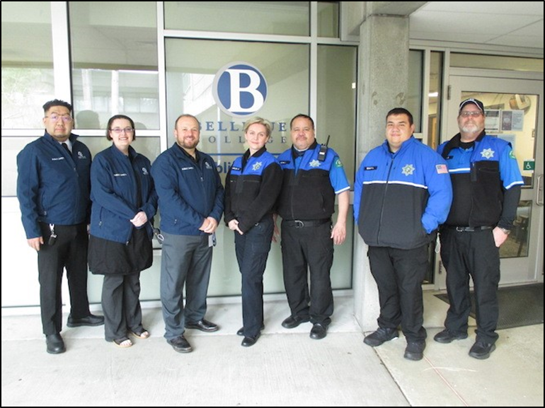 Seven public safety staff and officers wearing black and blue uniforms standing in front of the windows of the Public Safety Office.