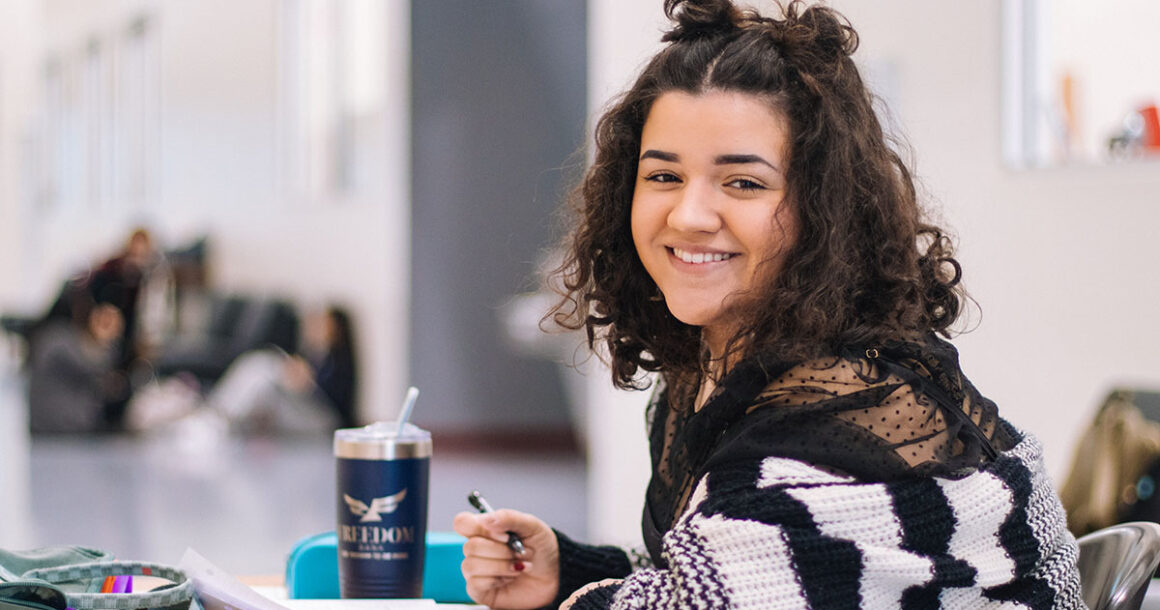 Student smiling sitting at a table with book open and pen in hand.