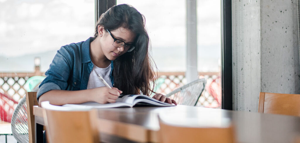 A student fills out workbook exercises at a table near a window.