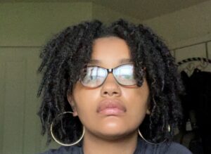 A photo of Madison looking at the camera with short curly brown hair wearing glasses and gold hoops earrings