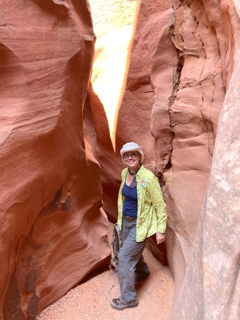 Mandie smiles, standing surrounded by reddish brown canyon walls. She wears a yellow jacket, grey pants, and a white hat while carrying a backpack in one hand.