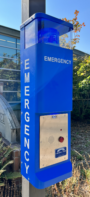 A blue emergency phone with the word "Emergency" written over the side features a large light on the top, a big red button, and speaker grate.