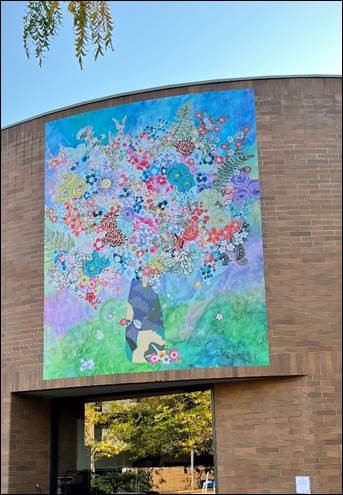 A bright mural of flowers and plants covers the bricks of a building in soft colors that evoke nature and life. The image sits above a walkway and below a clear blue sky.