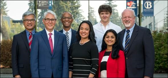 The Board of Trustees and Interim President Locke stand together smiling, campus buildings and trees in the background.