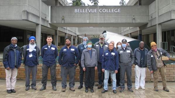 Warehouse and Maintenance crew pose for a picture in front of the fountain and "Bellevue College" sign.