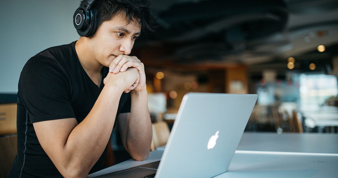 Student sitting at a table with headphones in front of a mac laptop.