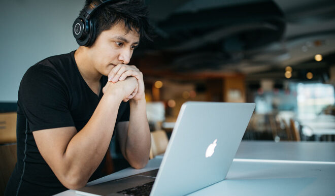 Student sitting at a table with headphones in front of a mac laptop.