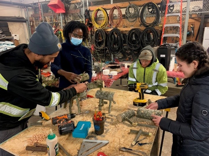 Four individuals working in a workshop surrounding a table filled with various tools to make reindeer from pieces of trees.