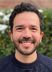 Felipe smiling, wearing a navy blue knit sweater with green foliage behind