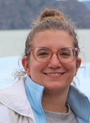 Maria smiling, with pink glasses and a white jacket, on the beach with water, sand, and mountains behind