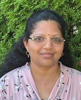 Mathi smiles wearing glasses and round earrings, with green bush leaves in the background