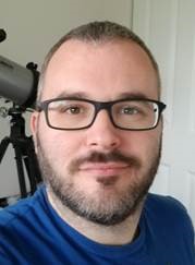 Dr. Tim smiling and wearing a dark blue shirt and black glasses, with a telescope in the background