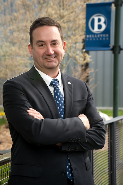 On campus, David May smiles with arms crossed, wearing a black suit with a blue BC logo print tie.