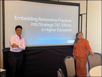 Two people stand smiling on either side of a projector screen displaying a slide that reads: "Embedding Restorative Practices into Strategic DEI Efforts in Higher Education."