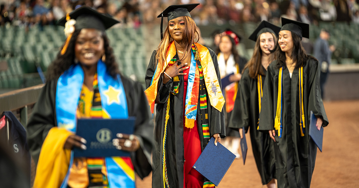 Bellevue College graduates wearing graduation gowns and caps walk across a field carrying diplomas.