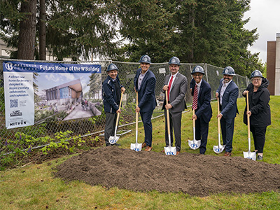 Six people wearing hard hats stand with shovels near a pile of dirt outside.