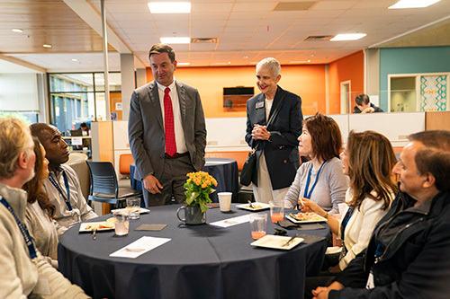 Two people standing chat with a group of people at a table. 
