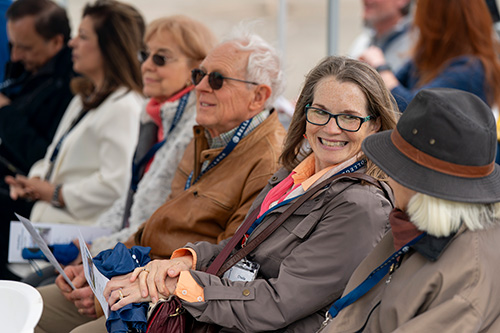 People sitting in a row of chairs, one one smiles as she looks at person sitting next to her.