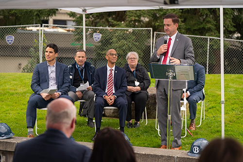 Man stands at podium with microphone under white tend with four other speakers seated behind him.