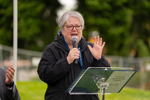 Woman holding microphone and holding up one hand while speaking.