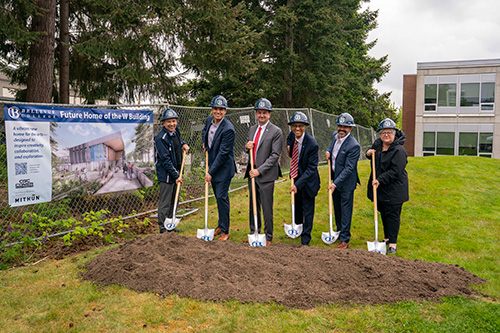 Six people wearing hard hats pose with shovels in a mound of dirt. 