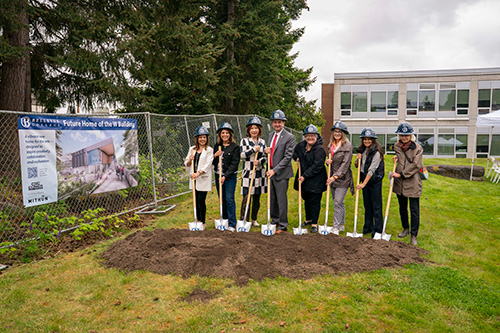 Eight people wearing hard hats pose with shovels in dirt with a fence and sign to the left.