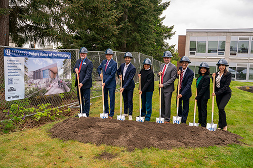 Eight people wearing hard hats pose with shovels in dirt with a fence and sign to the left.