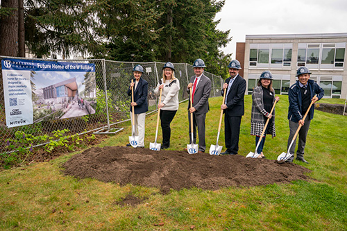 Six people wearing hard hats pose with shovels in dirt with a fence and sign to the left.